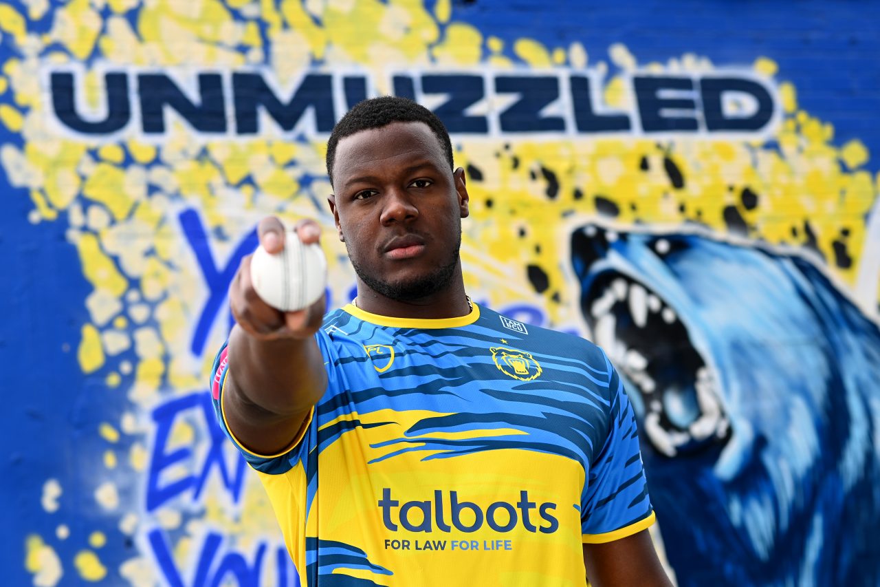 Cricketer wearing blue and yellow cricket gear holds out a cricket ball towards the viewer. Unmuzzled written on the wall behind him with a snarling bear to his right.