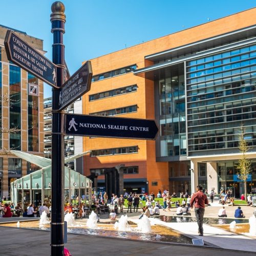A sign post in Central Square, Brindleyplace with the sun shining and the fountains on with people enjoying the afternoon.