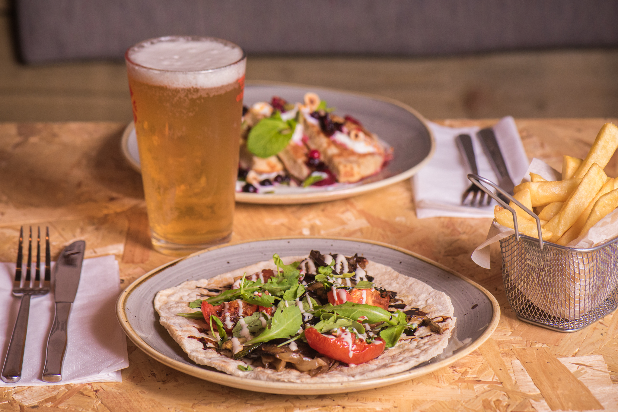 A table set for two, with two mains and a pint of beer. One main is savoury with flat bread salad and mushrooms and the other is sweet with berries, nuts and seads. 