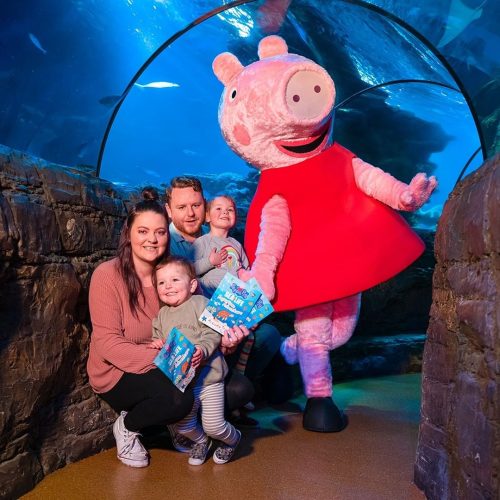 Peppe Pig with a mom, dad and two children in the SEA LIFE Centre Ocean Tunnel with the aquarium and fish visible behind them.