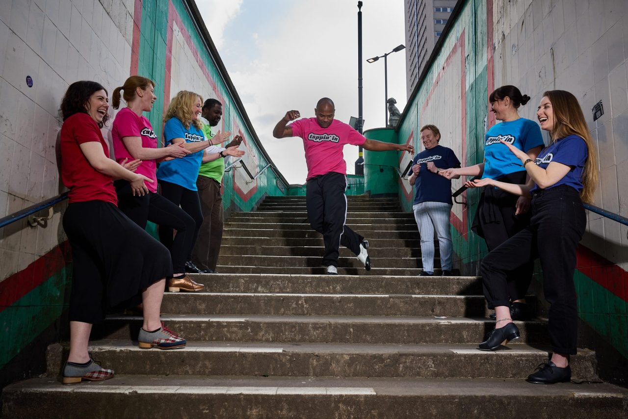 A diverse group of people stand in a row on either side of a concrete stair as one man dances in the middle.