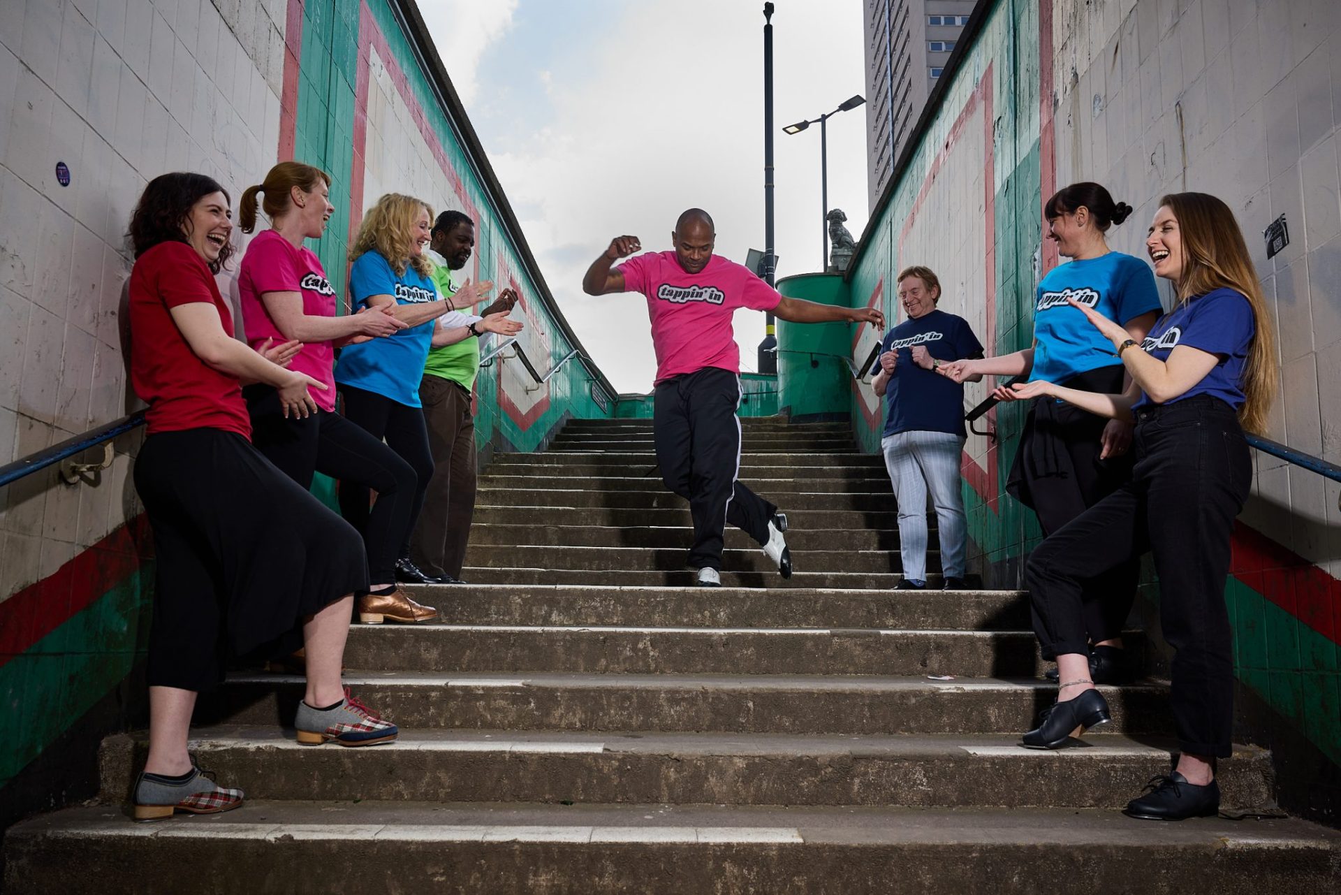 A diverse group of people stand in a row on either side of a concrete stair as one man dances in the middle.