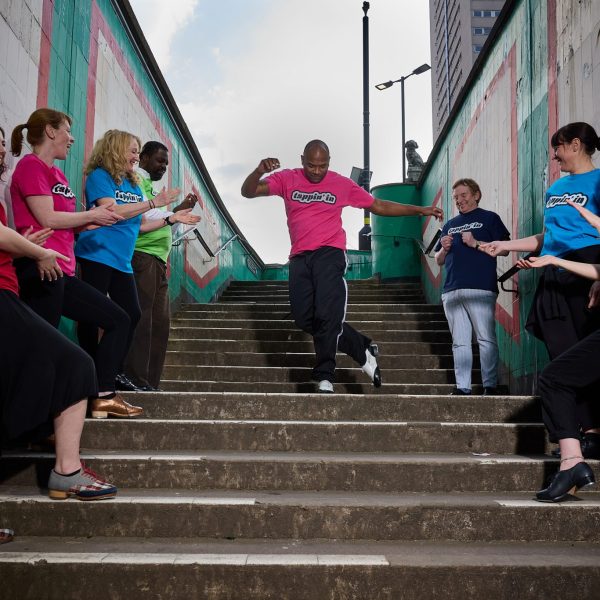 A diverse group of people stand in a row on either side of a concrete stair as one man dances in the middle.