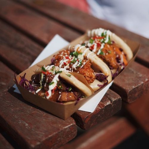 Three boa buns sit side by side in a brown cardboard container on a brown wooden picnic table.