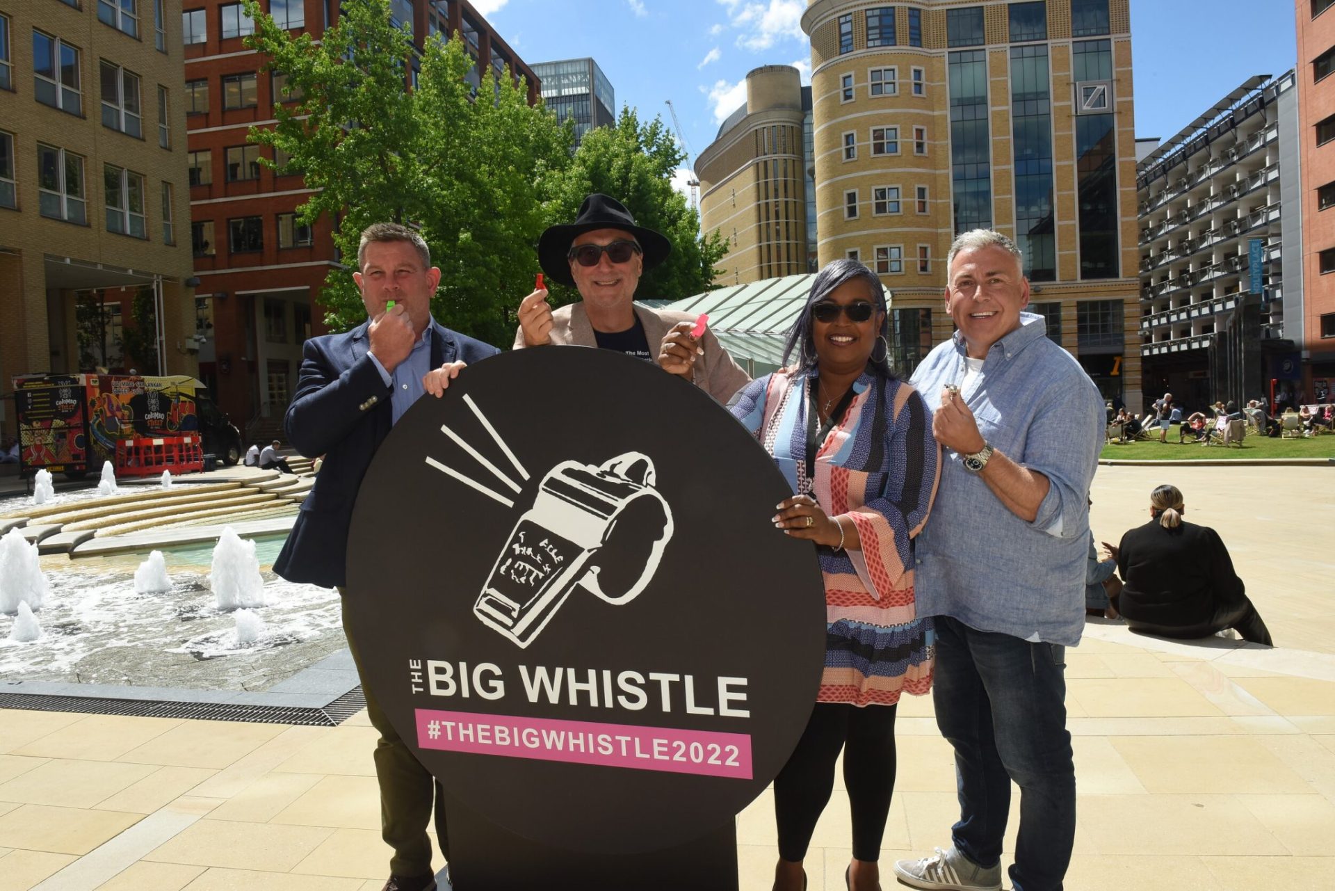 a group of four people stand behind a stand black circular stand with a shite whistle on it with the hastag the Big whistle 2022