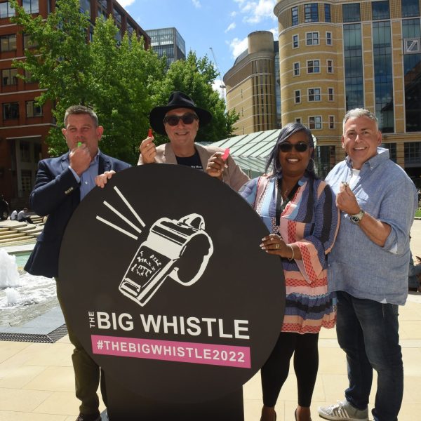 a group of four people stand behind a stand black circular stand with a shite whistle on it with the hastag the Big whistle 2022