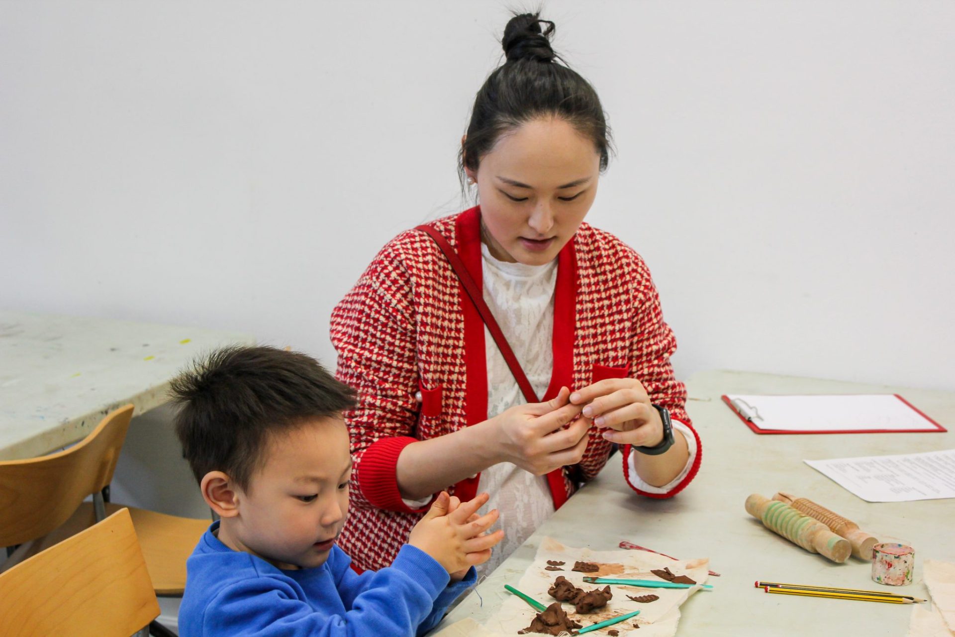 A mother in a red ad white cardigan and son sit at a table creating art made of clay.