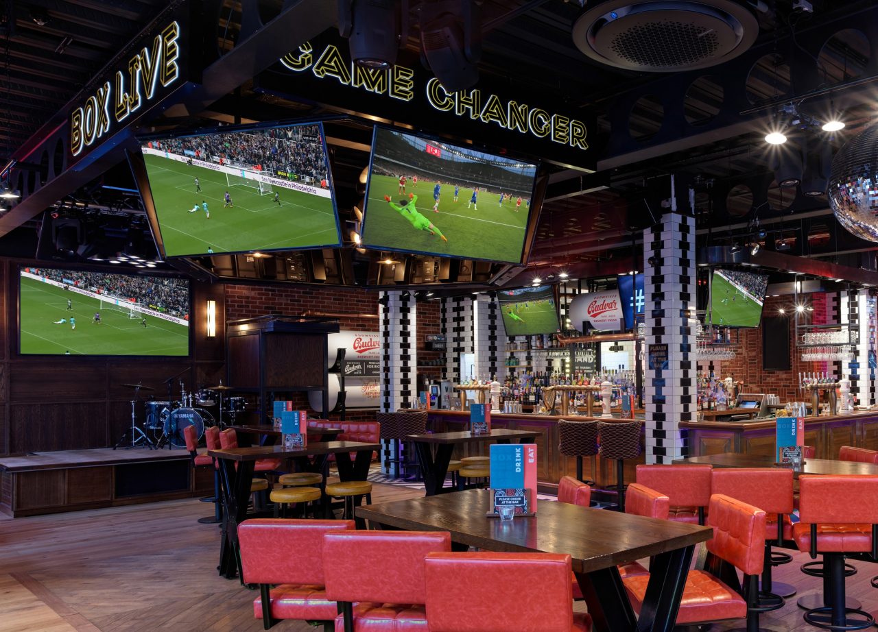 The interior of a sports bar, large TVs hanging from the ceiling showing a football match, with oarnge bar height chairs around tables.