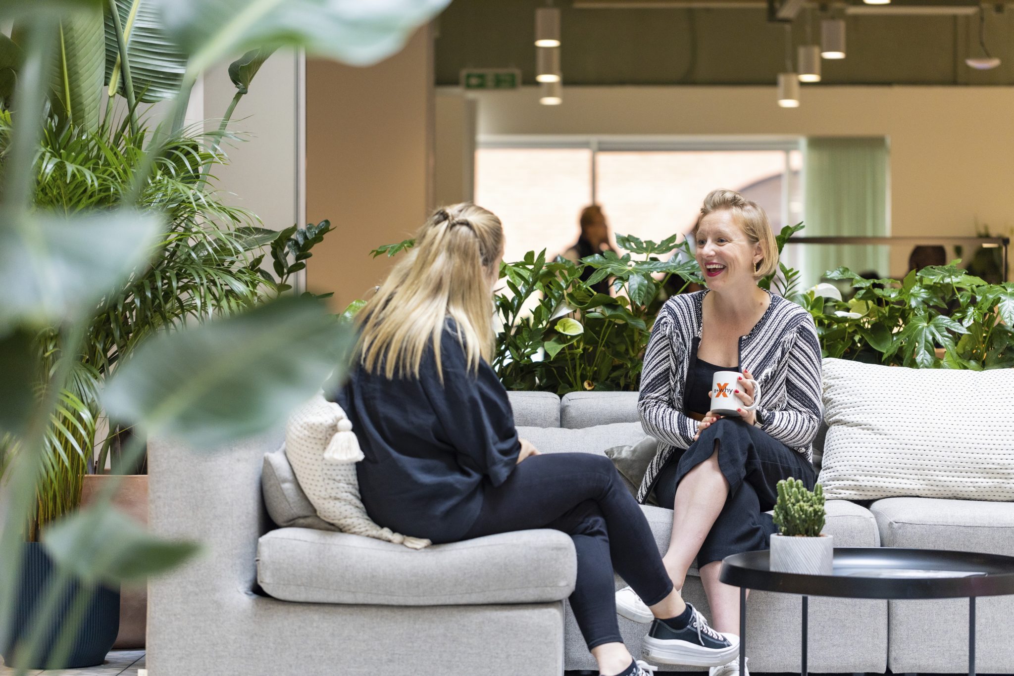 Two woman sit facing eachother on a beige couch, drinking coffee and chatting in the x+why atrium.